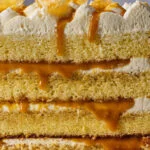 Close-up of a layered yellow sponge cake with whipped cream, caramel drizzle, and almond shards on top.