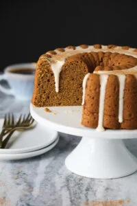 Pumpkin cake with cream glaze on a white stand, a slice removed showing moist interior, coffee cup in the blurred background.