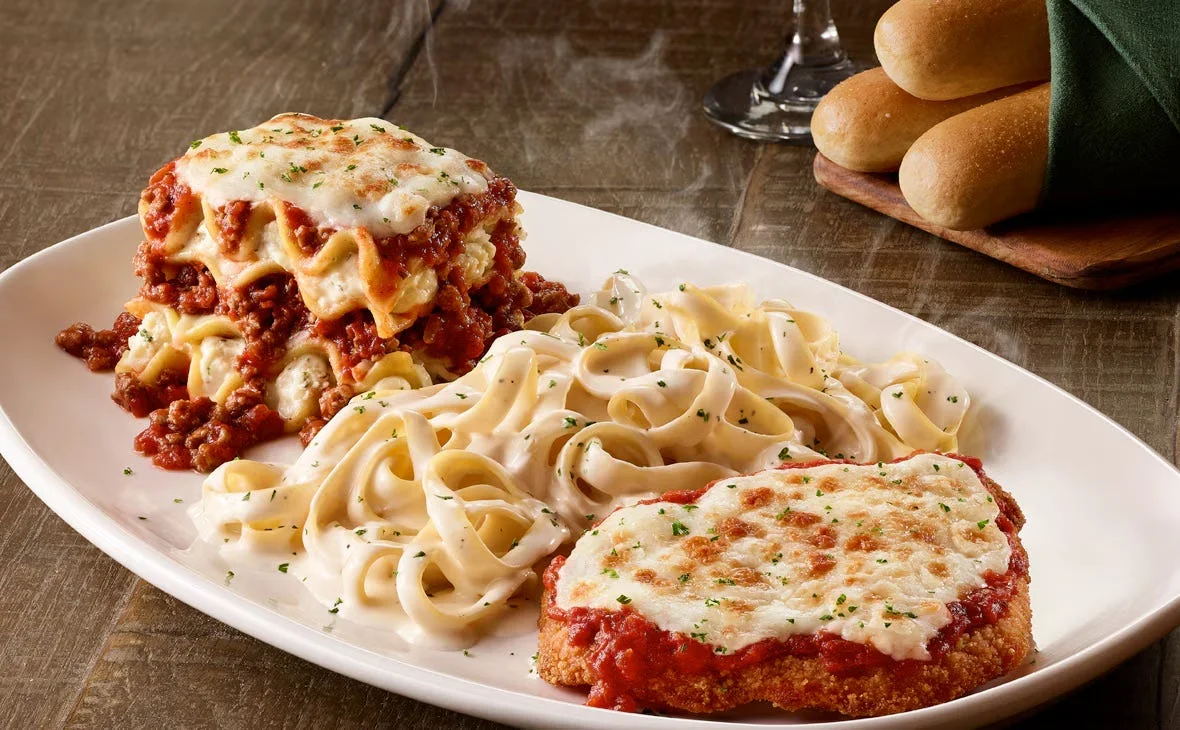 A plate of lasagna, chicken parmesan, and fettuccine alfredo with herb garnish and breadsticks on the side.