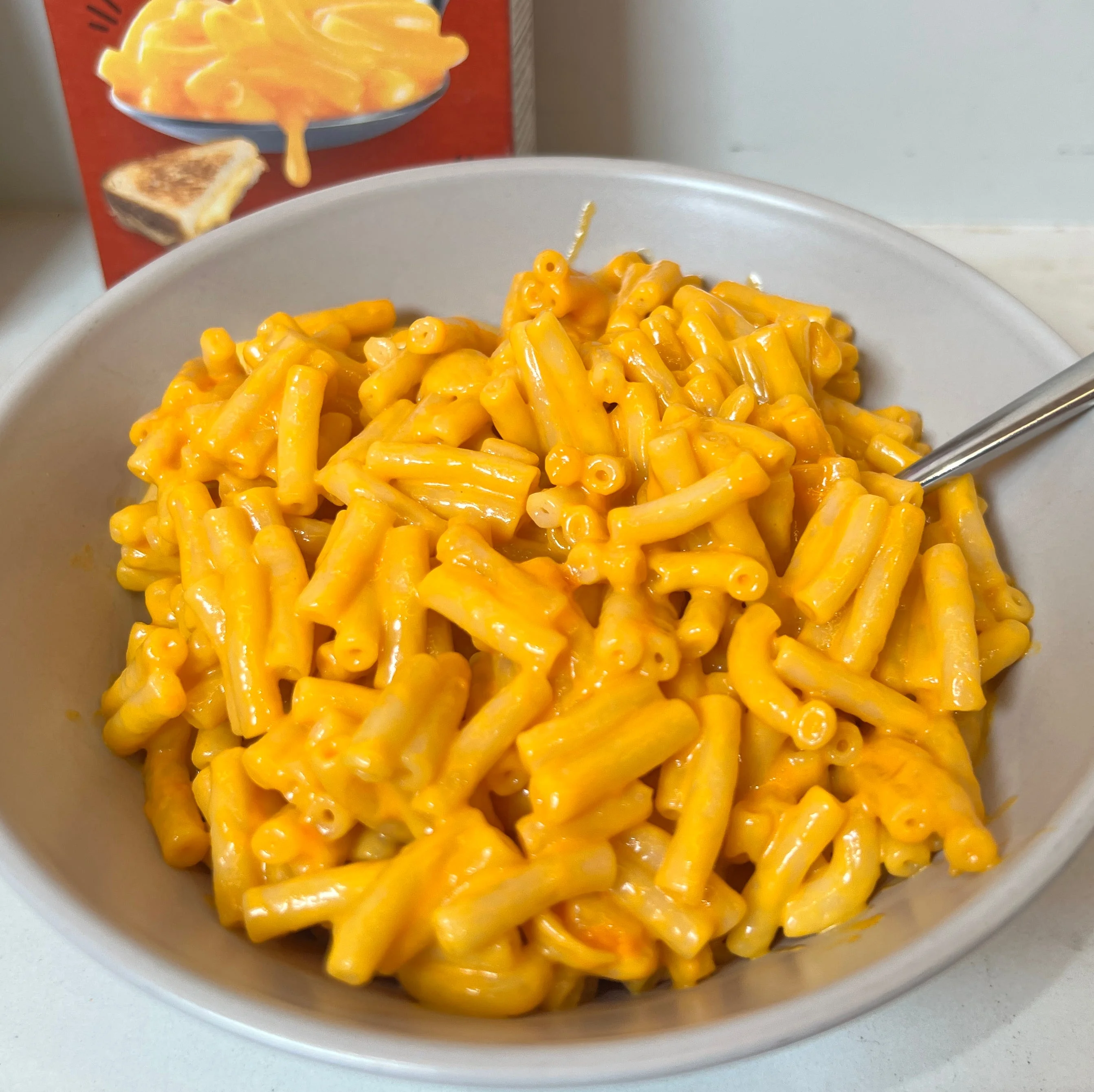 Bowl of cheesy macaroni pasta with a spoon, set on a white surface next to packaging. Bowl of cheesy macaroni pasta with a spoon, set on a white surface next to packaging.