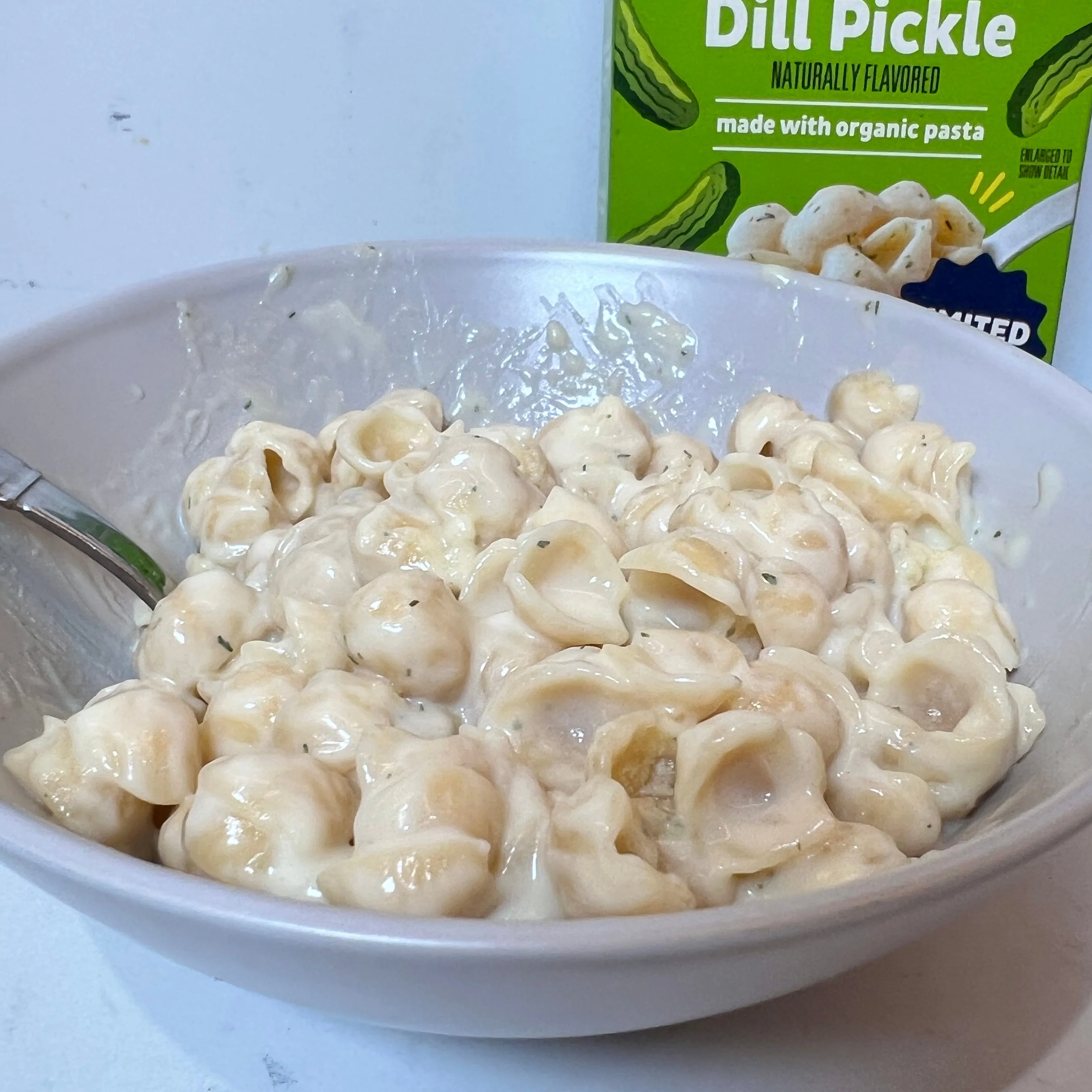 Bowl of creamy dill pickle-flavored pasta with packaging of similar products in the background. Bowl of creamy dill pickle-flavored pasta with packaging of similar products in the background.